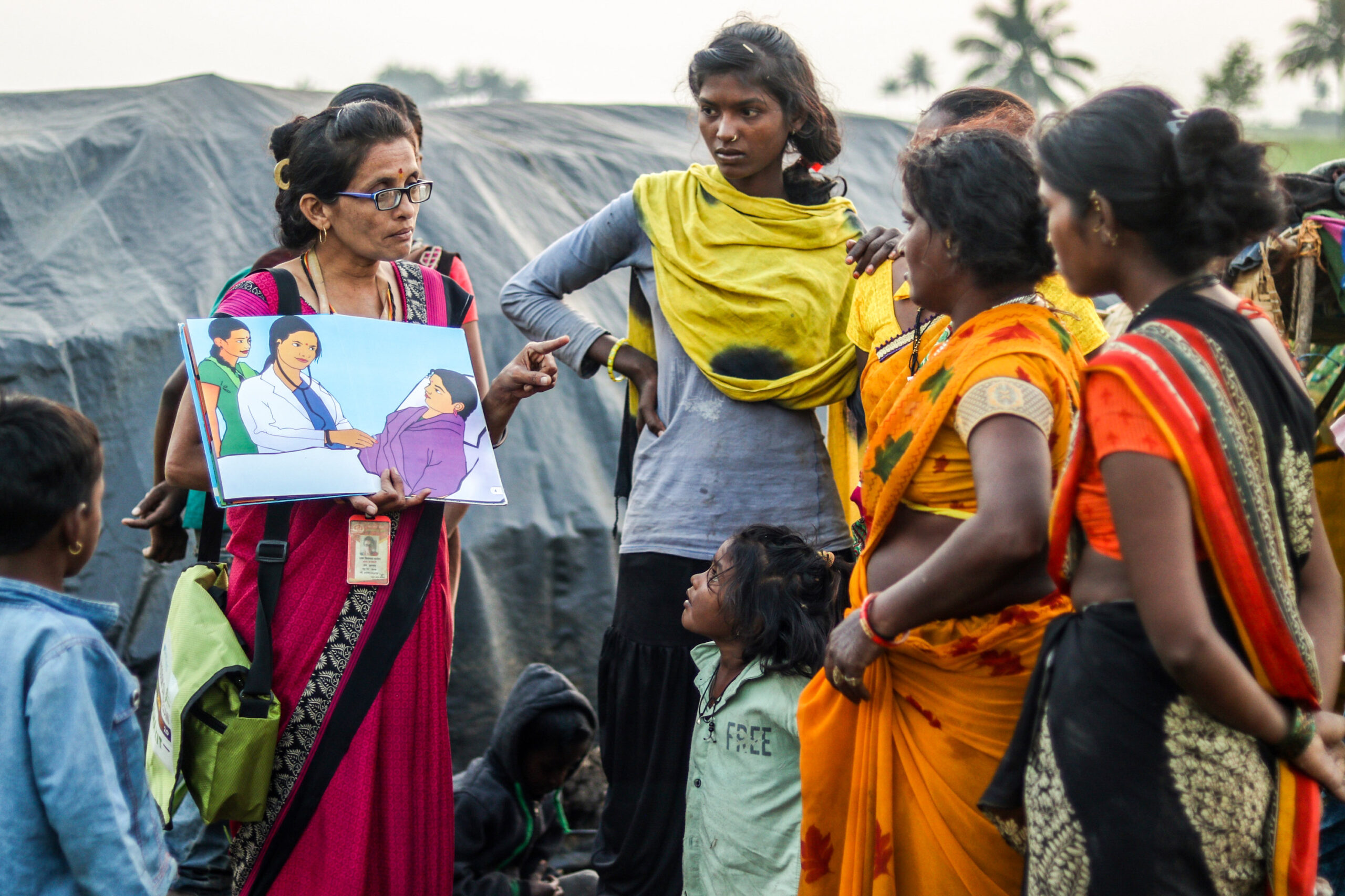 Maya Patil standing outdoors in a group of young women and children and holding up an illustrated book