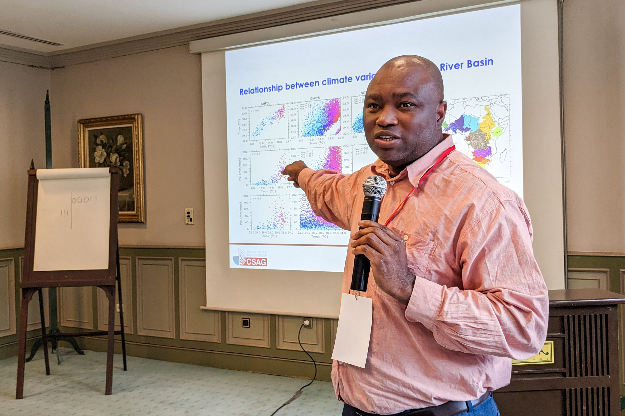 Professor Abiodun holds a microphone and points back toward climate statistics projected on the screen behind him