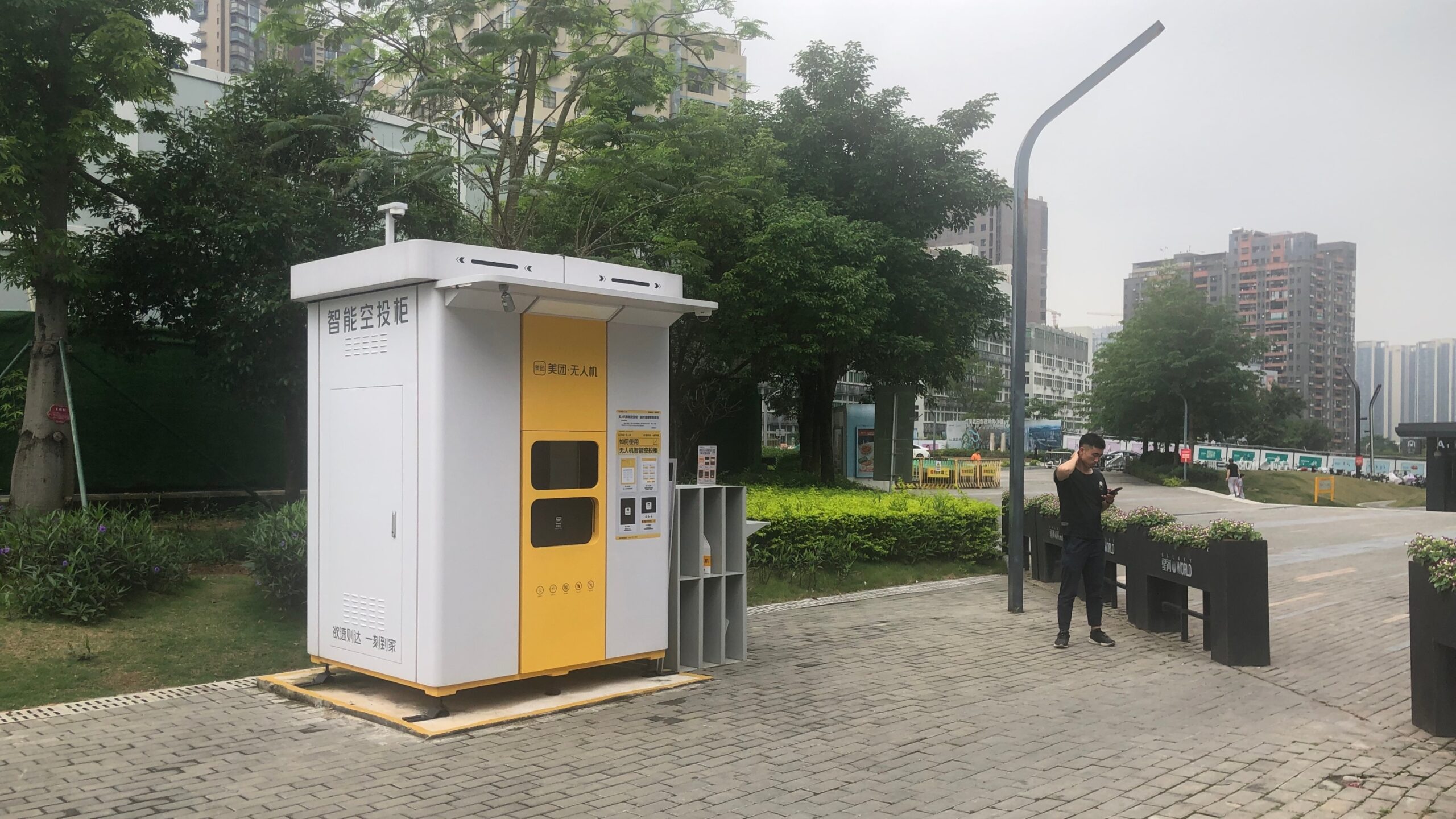 A yellow-and-white Meituan pickup kiosk in front of trees. A man is standing nearby.