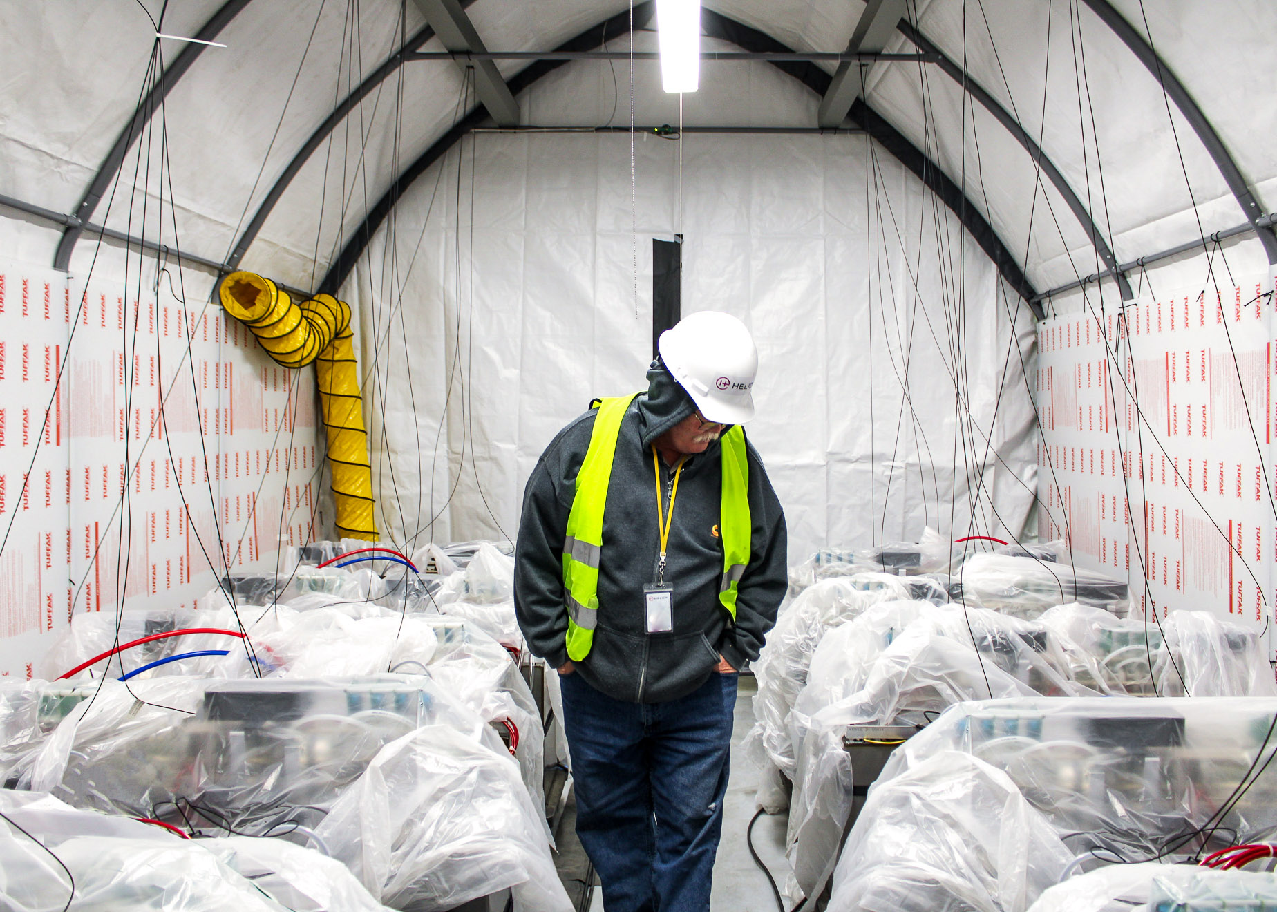 a worker in a Helion hard hat walked down an aisle surrounded by machinery covered in plastic sheeting
