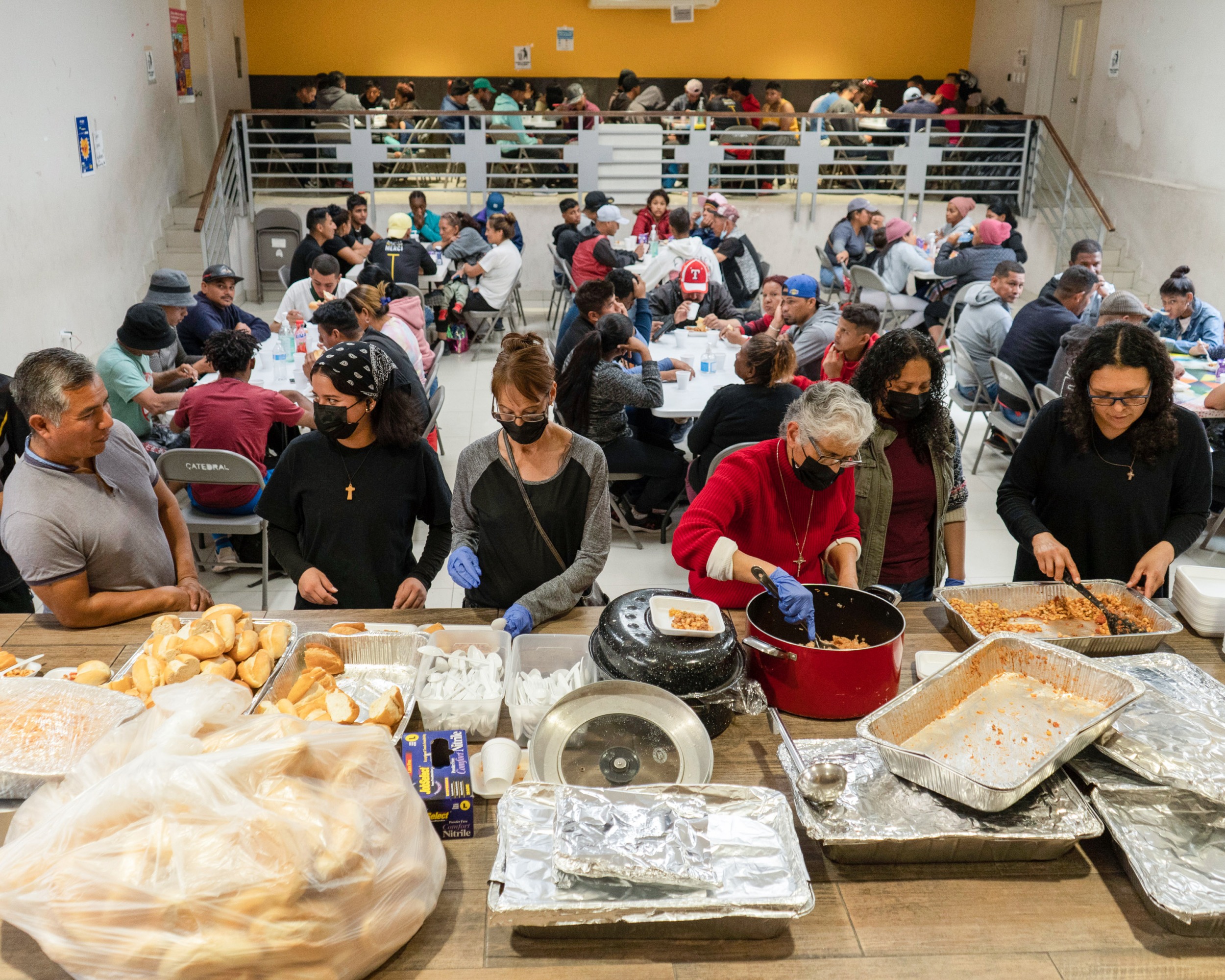 In the foreground people help themselves from aluminum trays of food while others can be seen sitting at tables eating in the background.