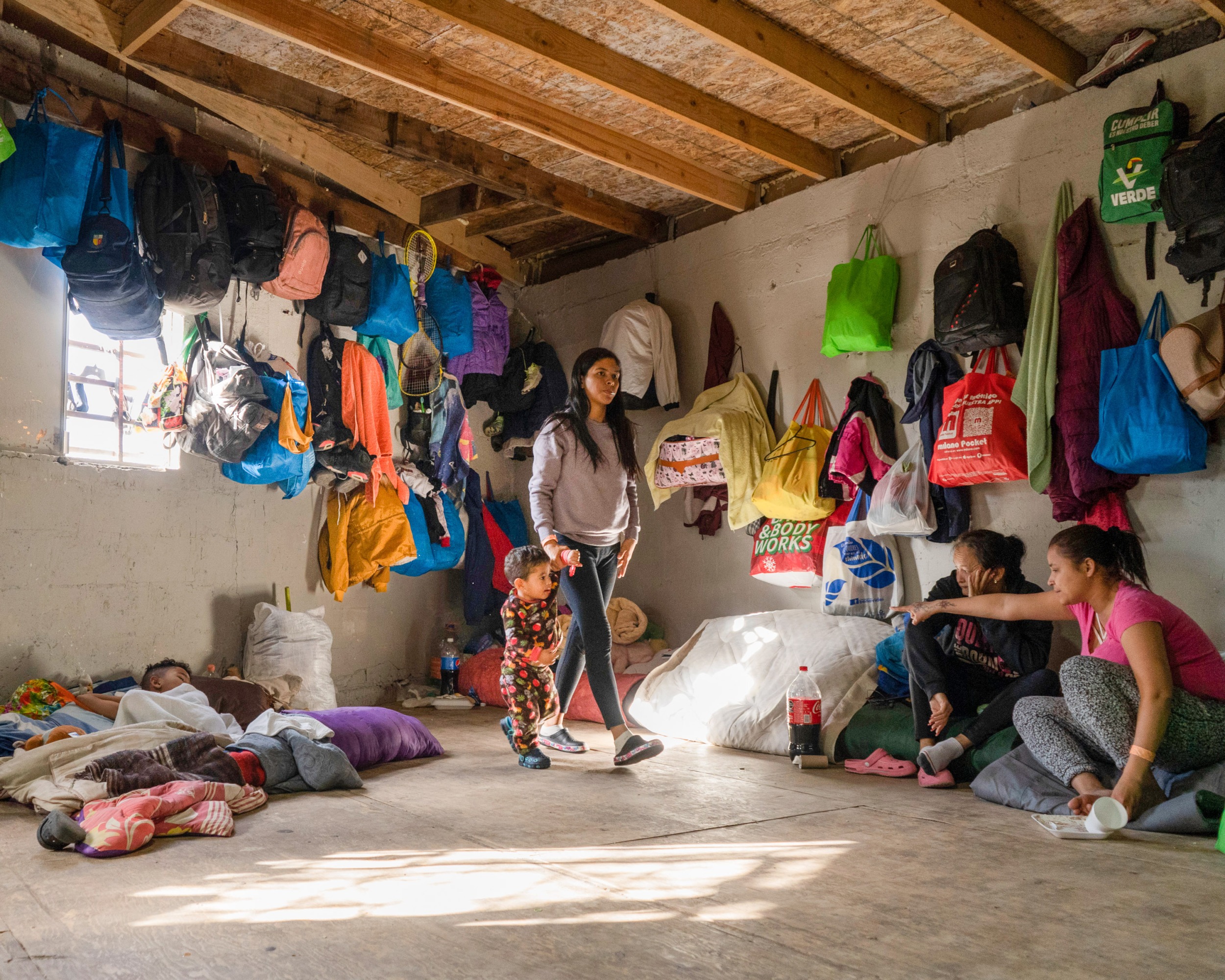 a woman leads her son through a room where people and belongings are up against a wall