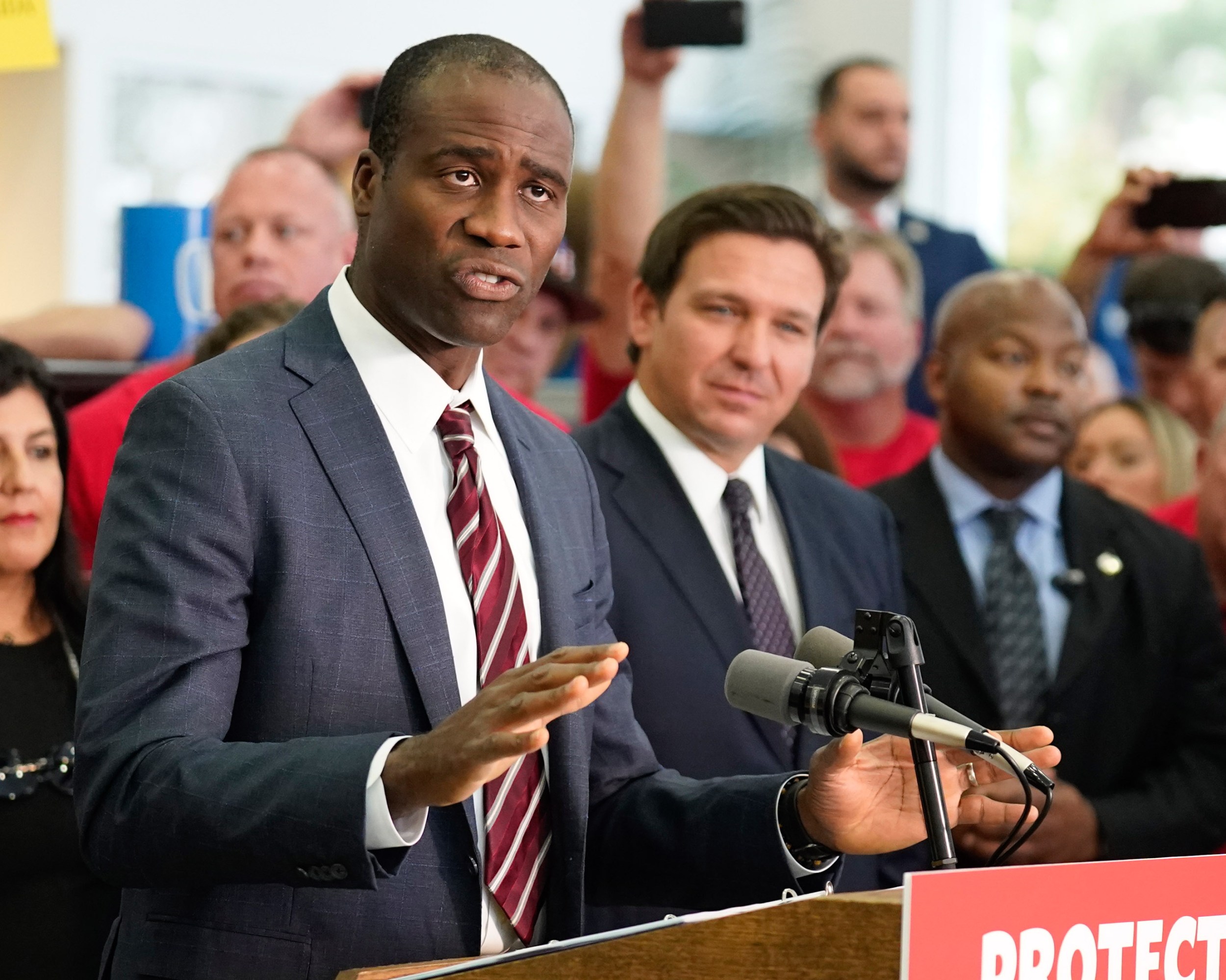 Florida Surgeon Gen. Dr. Joseph A. Ladapo speaking at a podium with Ron DeSantis behind him