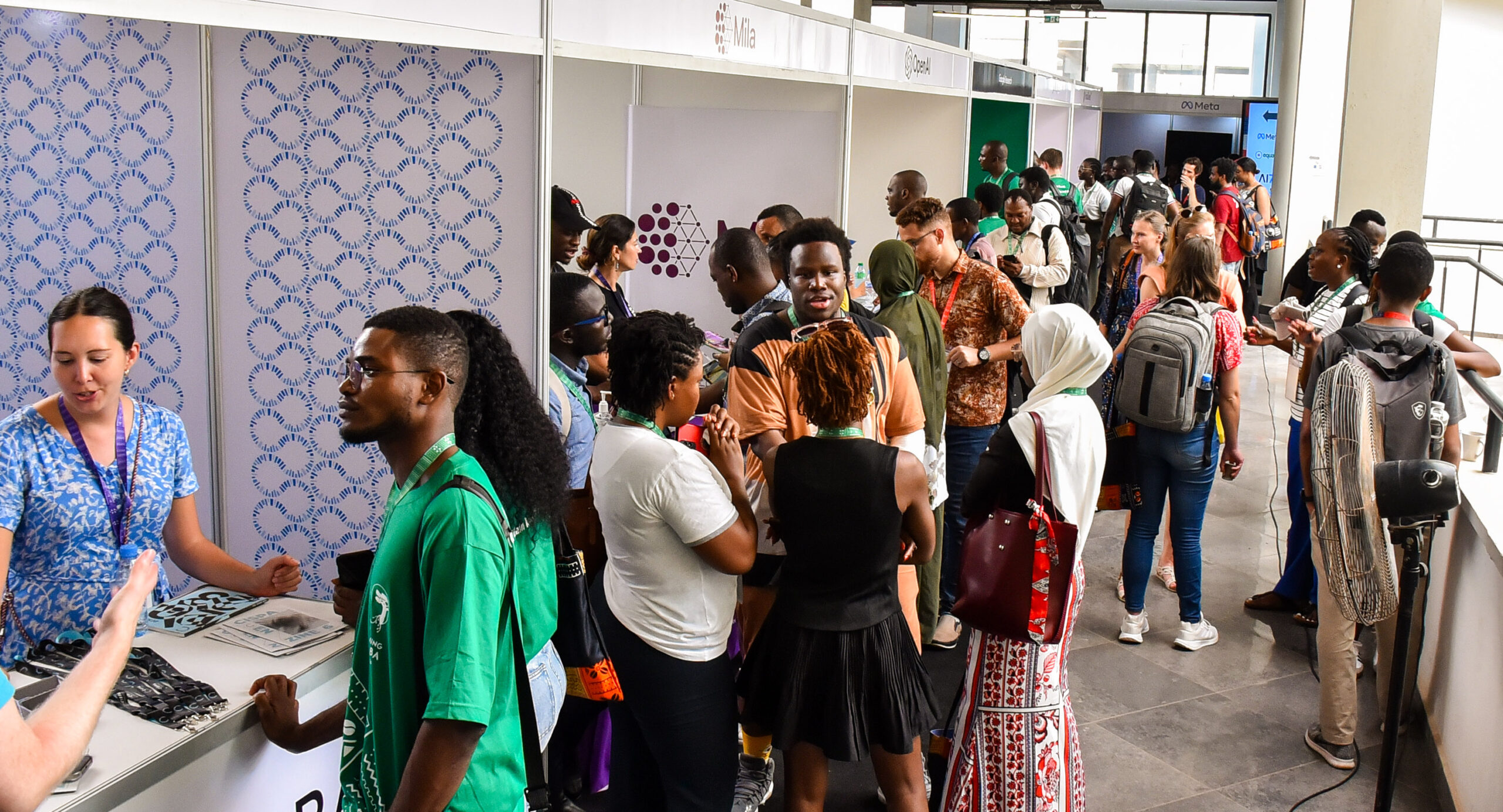 crowd of attendees mingle around display booths at Deep Learning Indaba 2024. Booth signs for Mila, Meta and OpenAI can be seen in the frame.