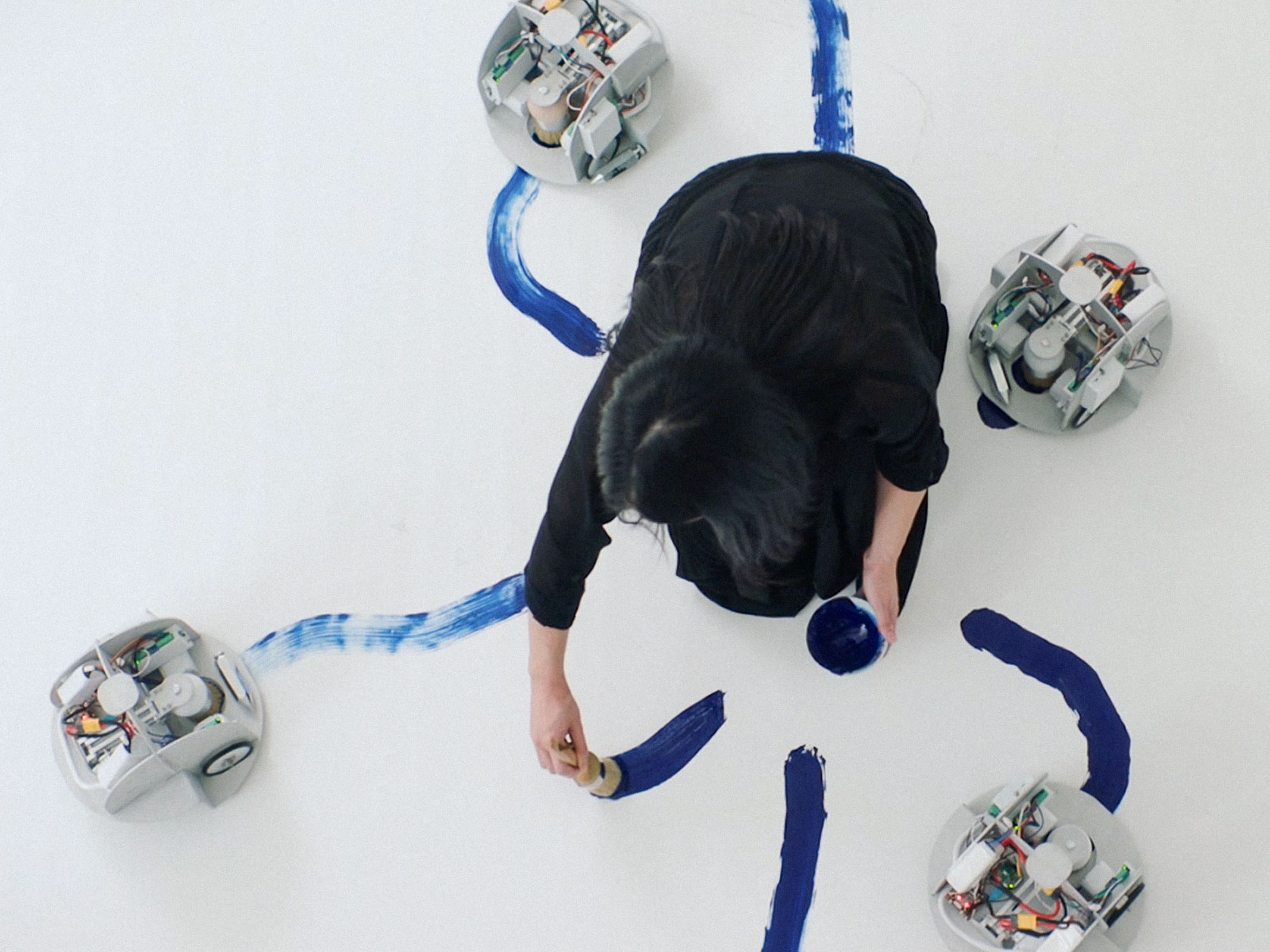 artist from overhead kneeling on a surface making blue paint swipes with 4 robots