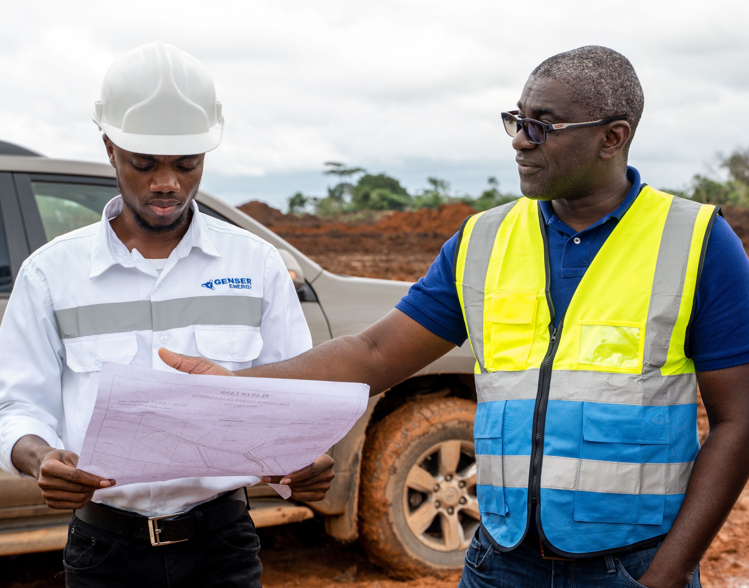 two men in safety vests standing in a muddy area looking over a document