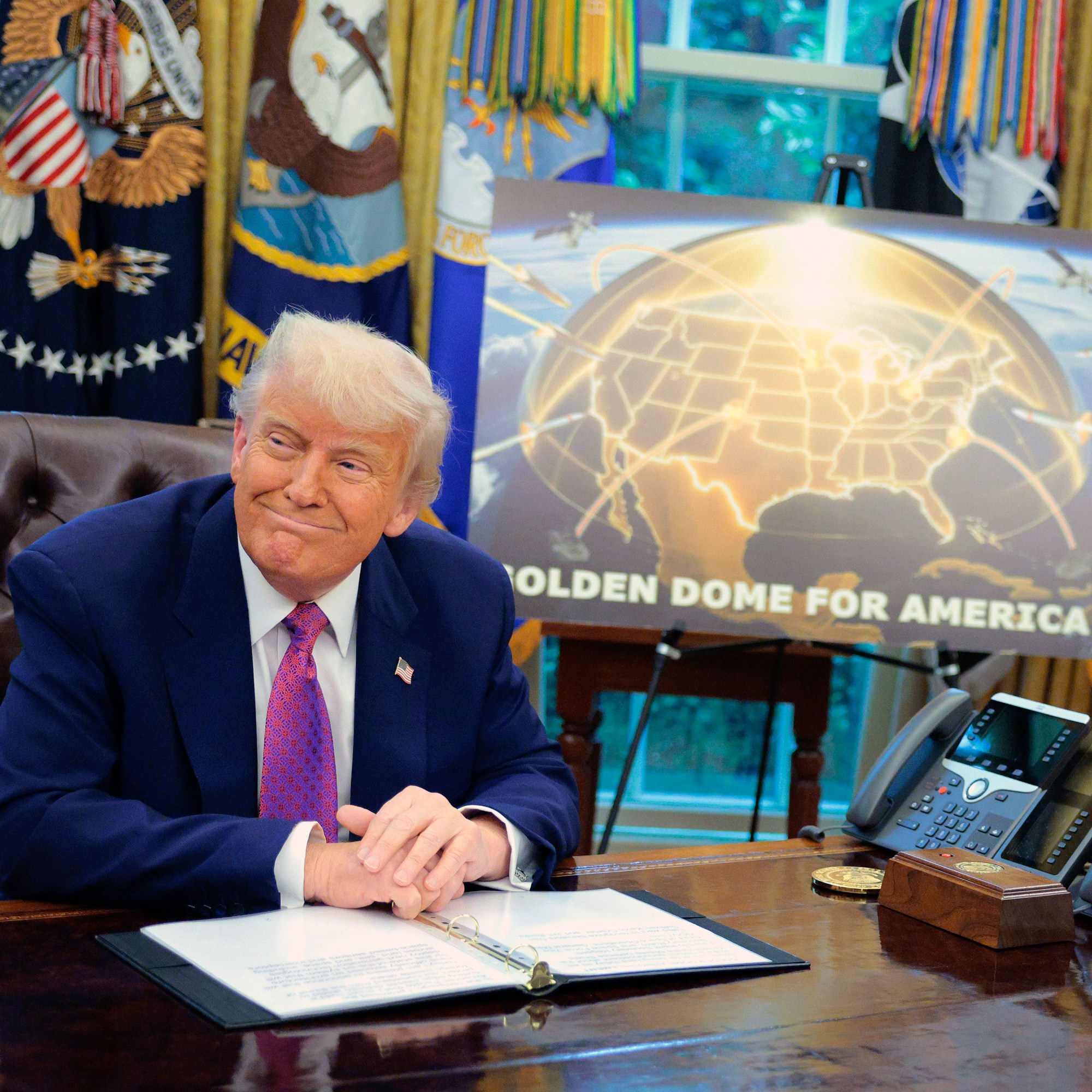 Trump at the desk the Oval Office with a sign reading "Golden Dome for America" on a display stand behind him